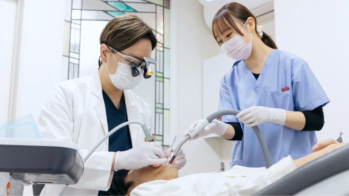 歯科医師と歯科衛生士が患者の歯の治療を行っている様子。歯科用ルーペと吸引装置を使用している。