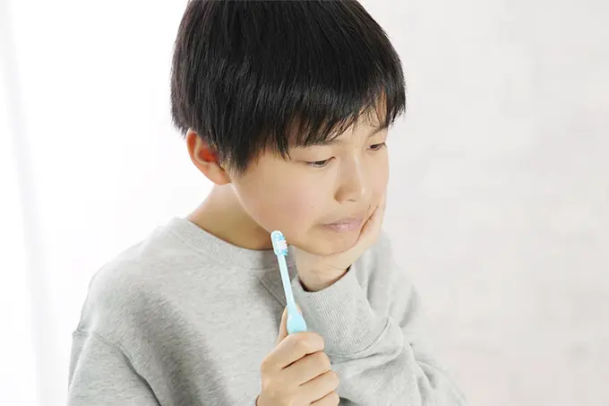 歯ブラシを持ちながら頬を押さえ、歯の痛みを感じている少年。虫歯や歯肉炎を示唆する表情。