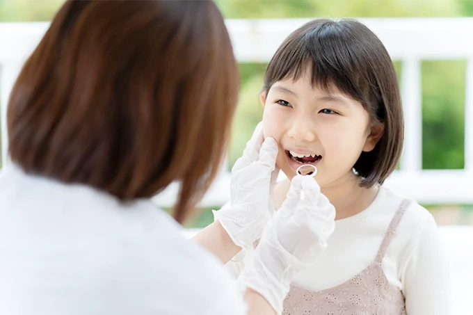 歯科医が女の子の口の中を診察している様子。女の子は笑顔で、歯科医は白い手袋を着用している。屋外の光が差し込む明るい診察風景。