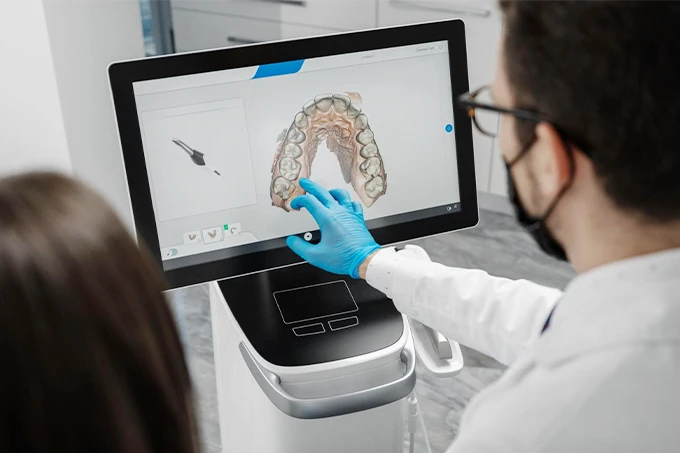 歯科医がタッチパネルモニターに表示された上顎の歯列3D画像を指しながら、患者に説明している様子。抜歯（難抜歯）に関するカウンセリングの一場面。