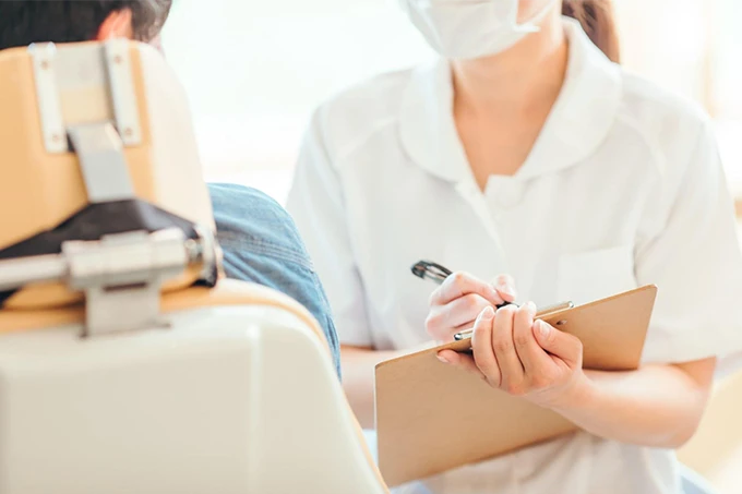 白衣を着た歯科スタッフが、診療チェアに座る患者と対面しながら問診票を記入している様子。初診時のカウンセリングシーン。
