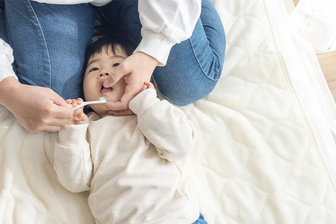 保護者が膝の上に子どもを寝かせ、仕上げ磨きをしている様子。家庭での歯みがき習慣の一場面。