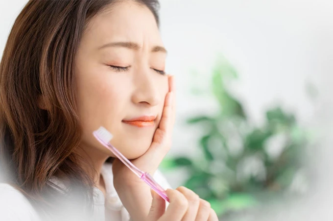 歯ブラシを持ちながら頬を押さえて歯の痛みに悩む女性。虫歯や歯周病の症状をイメージさせる。